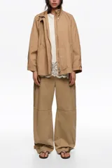Gabardina corta de línea A y tejido técnico color tostado, con cuello alto, cierre frontal con cremallera metálica, mangas largas con trabillas ajustables y bolsillos de vivo.