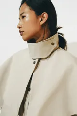Gabardina larga de corte recto y diseño minimalista en color beige, con cuello solapa, manga larga y cierre frontal con botones. Cuenta con una capa desmontable en el mismo tejido y una solapa decorativa en el pecho.