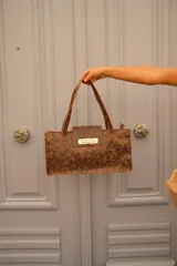 Cartera tipo tote bag de formato rectangular y alargado, confeccionada en gamuza color marrón oscuro. Presenta doble asa de hombro, solapa frontal con cierre metálico giratorio en tono dorado y costuras visibles en los bordes.