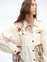 Chaqueta larga de diseño patchwork en tonos grises, con combinación de texturas que incluyen paneles de tela lisa, tela texturizada blanca y paneles con estampado floral en relieve. Presenta cierre frontal con botones, cuello camisero y bordes deshilachados.