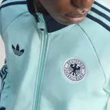 Campera deportiva Adidas Originals de la selección de Alemania, color celeste con detalles en azul marino en cuello, puños y las icónicas tres tiras en las mangas. Presenta cierre frontal, logo del Trifolio y escudo de la federación alemana de fútbol en el pecho.