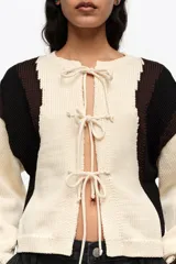 Cardigan de punto con diseño de bloques de color en blanco, negro y marrón, escote redondo y cierre frontal con múltiples lazos de tela.