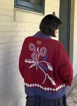 Saco tejido tipo jacquard color bordó con diseño floral en la espalda, cuello alto y cierre frontal de aluminio. Presenta detalles en azul y blanco en el cuello, puños y ruedo, con un patrón geométrico de rombos en la parte inferior.