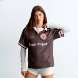 Camiseta de fútbol Nude Project de corte oversize, color marrón con detalles en rosado, cuello polo y escote en V. Cuenta con estampado frontal de la marca, frase bordada y escudo circular con una rosa en el pecho.