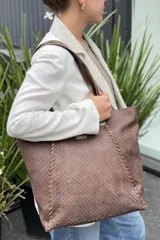 Bolso grande tipo tote color beige con textura tejida y costuras decorativas en los laterales. Tiene doble asa de hombro y logo de la marca en el frente. Interior forrado con bolsillo con cierre y portacelular.