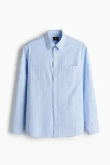 Camisa de popelina de algodón con diseño de rayas verticales celestes y blancas, corte holgado, cuello clásico, bolsillo frontal en el pecho y mangas largas con puños abotonados.