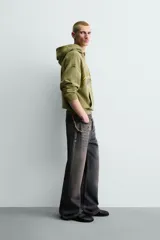 Sudadera con capucha de corte relajado en color verde oliva con efecto lavado. Presenta un bolsillo frontal tipo canguro y un estampado gráfico en el pecho con letras en tonos amarillos y negros. Cuenta con puños y bajo acanalados.