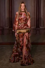 Vestido largo de mujer estilo boho en tono marrón, con estampado paisley y floral, cuello alto, abertura frontal en el escote y mangas largas abullonadas. Cuenta con detalles de aplicaciones de pedrería y flecos en la cintura, y falda larga con tajo lateral.