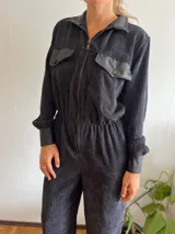 Mono largo de pana color negro, con cuello camisero y cierre frontal con cremallera. Presenta bolsillos con solapa de cuero en el pecho y puños de cuero.