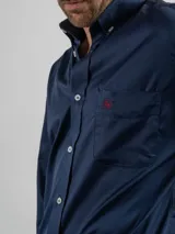 Camisa de corte regular color azul marino, confeccionada en algodón. Presenta cuello con botones, cierre frontal abotonado y un bolsillo en el pecho con el logo de la marca bordado en color rojo.