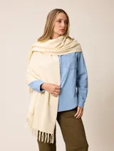 Pashmina de textura suave y color crema, con diseño liso y terminaciones en flecos en los extremos. Incluye una etiqueta decorativa en una de las esquinas.