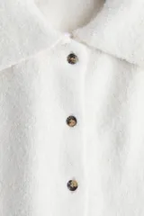 Saco de punto suave color crema, con cuello camisero y cierre frontal de botones marrones. Presenta hombros caídos y mangas holgadas por debajo del codo.