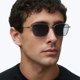 Lentes de sol con montura de acero inoxidable en color gris mate y lentes rectangulares polarizados en color negro. Presentan un diseño minimalista de líneas limpias con terminaciones de acetato en las patillas.