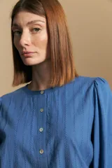 Camisa de algodón blanca con delicadas líneas bordadas verticales y textura calada. Presenta escote redondo con cierre frontal de botones, mangas largas con leve volumen en los hombros y puños elásticos.