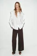 Camisa blanca de manga larga con un top de sudadera gris claro superpuesto, que incluye capucha ajustable con cordones. El bajo de la camisa tiene aberturas laterales.