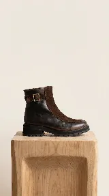 Bota urbana de caña media, confeccionada en una combinación de cuero gastado y gamuza. Presenta una solapa frontal en gamuza, cierre lateral y una tira decorativa con hebilla metálica en el tobillo. Cuenta con una suela de goma robusta y dentada.