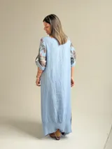 Vestido largo celeste con gran estampado floral marrón en el lateral derecho, de corte holgado, mangas 3/4 con trabilla y dobladillo irregular con flecos desflecados. Incluye un bolsillo aplicado en el frente.
