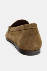 Mocasín de mujer confeccionado en serraje color marrón, con detalle de antifaz y borlas en el empeine. Presenta punta redonda y suela plana.