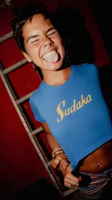 Remera tipo crop top de color azul con estampa frontal en color amarillo que dice 'Sudaka'. Presenta un ajuste entallado y cuello redondo.