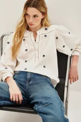 Camisa de algodón color crudo con bordados florales pequeños en azul marino. Presenta mangas largas abullonadas, cuello redondo y una abertura frontal con botones y detalles de encaje calado en la pechera y hombros.