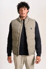 Chaleco puffer de corte regular, color beige, con cierre frontal y cuello alto. Presenta un diseño acolchado horizontal y logo bordado en el pecho.