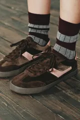 Zapatillas deportivas de estilo urbano, confeccionadas en una combinación de gamuza marrón oscuro y detalles en cuero rosado en el lateral y el talón. Presentan cierre mediante cordones al tono y suela de goma plana.