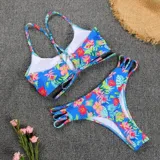 Bikini de dos piezas con estampado tropical de flores y hojas en tonos rosados, azules y verdes sobre fondo celeste. El corpiño es de diseño triangular con espalda cruzada, y la bombacha es cola less con tiras laterales decorativas.