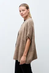 Capa tejida de punto en color beige, con diseño abierto y cierre frontal mediante broche metálico con detalles en cuero sintético negro. Presenta mangas cortas y textura suave.