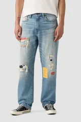 Jean denim de corte holgado, color azul claro, con efecto desgastado y roturas. Presenta parches de tela estampada en rojo con puntos blancos, estampado de vaca en blanco y negro, y un parche amarillo a cuadros.