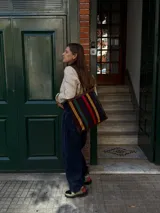 Cartera tipo tote de cuero con diseño de franjas verticales multicolores en tonos tierra, rojo y azul. Cuenta con correas trenzadas de cuero y cierre superior.