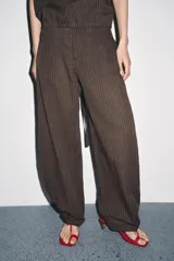 Pantalón de tiro medio confeccionado en hilatura con mezcla de lino a rayas verticales marrones y blancas. Presenta bolsillos laterales ocultos en costura, detalle de pliegues en el bajo y cierre frontal con cremallera, botón y ganchos metálicos.