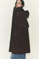 Chubasquero largo color marrón oscuro, con cuello subido y capucha ajustable con cordones. Tiene cierre frontal con cremallera oculta y botones a presión, y bolsillos delanteros con solapa y botón a presión oculto.