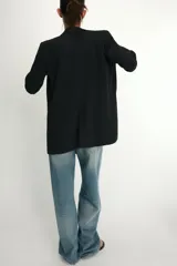 Blazer negro con cuello solapa con muescas, hombreras y forro interior a tono. Presenta manga por debajo del codo con detalle de drapeado y falsos bolsillos de vivo en el delantero, con cierre frontal de un solo botón dorado.