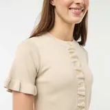 Camiseta de punto color beige con cuello redondo, cierre frontal de botones y mangas cortas con volantes. Presenta detalles de volantes en la botonera y en el ruedo.