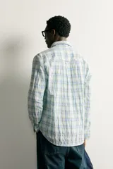 Camisa de corte regular fit con cuello abotonado, manga larga y bolsillo de parche en el pecho. Presenta un diseño a cuadros en tonos verdes y azules sobre fondo blanco, con un sutil efecto arrugado en el tejido.
