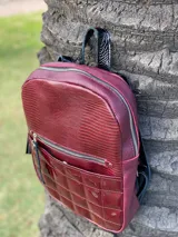 Mochila color bordó con textura tipo reptil en la parte superior y bolsillo frontal con diseño capitoneado en material charolado. Cuenta con cierre metálico y tiradores con flecos decorativos.