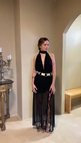 Vestido largo negro con escote profundo tipo halter y cuello alto. La falda presenta frunces y capas de microtul transparente. Se complementa con un cinturón ancho metálico de doble hebilla.