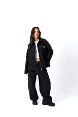 Camisa de gabardina negra de diseño unisex y corte holgado. Cuenta con un cierre frontal de doble camino, cuello camisero y detalles de estampa y bordado en el pecho con el texto 'Low Profile'.