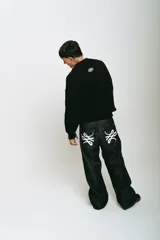 Pantalón de jean negro, corte baggy, confeccionado en denim crudo. Presenta un estampado serigrafiado en blanco con un diseño estilizado en ambos bolsillos traseros y un parche con logo en la cintura.