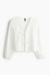 Blusa blanca de algodón con escote en V, frente abierto con cierre de tiras finas para anudar, mangas largas y puños elásticos.
