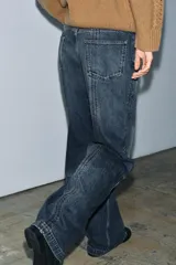 Jeans de mezclilla azul oscuro con efecto lavado, tiro medio y corte balloon (pierna ancha y holgada). Presenta cinco bolsillos y cremalleras metálicas decorativas que recorren la parte exterior de las piernas desde el muslo hasta el bajo.