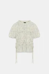 Blusa de manga corta abullonada con diseño a cuadros, cuello con volante y acabados elásticos. Cuenta con aberturas y lazos ajustables en los laterales.