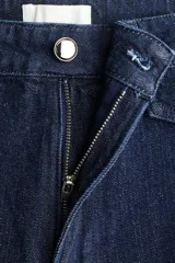 Pantalón de jean de denim azul oscuro, de tiro alto y piernas amplias con corte holgado. Presenta solapas de bolsillo decorativas con botón en el frente y costuras verticales visibles a lo largo de las piernas.