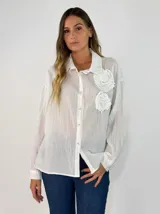 Camisa blanca de corte oversize y tejido semitransparente, con cuello clásico, cierre frontal de botones y detalle de flores aplicadas en el pecho.