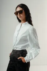 Camisa blanca de algodón con cuello alto tipo gorguera, cierre frontal con botones ocultos y mangas largas con puños elásticos.