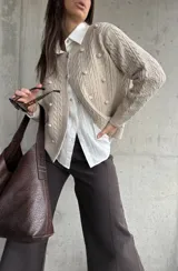 Cardigan de punto color beige con diseño de trenzas y pompones decorativos. Presenta cuello redondo, cierre frontal con botones y mangas largas.