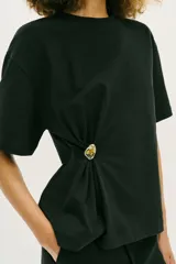 Camiseta de algodón compacto color negro, con cuello redondo, manga corta y detalle de aplique metálico en el frente que genera un ligero fruncido lateral.