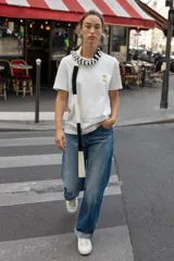 Camiseta negra de algodón, corte regular, con cuello redondo ribeteado en blanco y un bolsillo frontal con un broche metálico decorativo dorado.