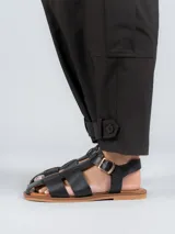 Sandalias planas de cuero negro estilo pescador, con tiras anchas entrelazadas en el empeine, suela marrón y tira ajustable con hebilla metálica en el tobillo.