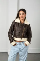 Chaqueta estilo aviador de cuero sintético en tono marrón desgastado, con cuello, puños y ruedo forrados en corderito sintético color crema. Presenta cierre frontal cruzado, bolsillos con solapa y cinturón ajustable en la cintura.